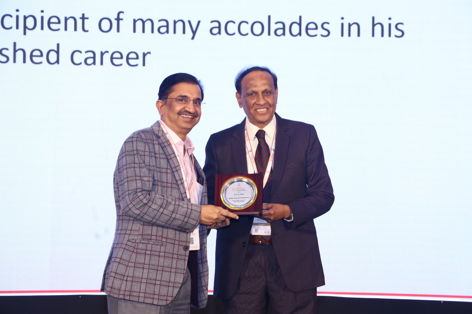 Two men smiling at the camera during a plaque handover on stage, both holding a framed Lifetime Achievement Award plaque.